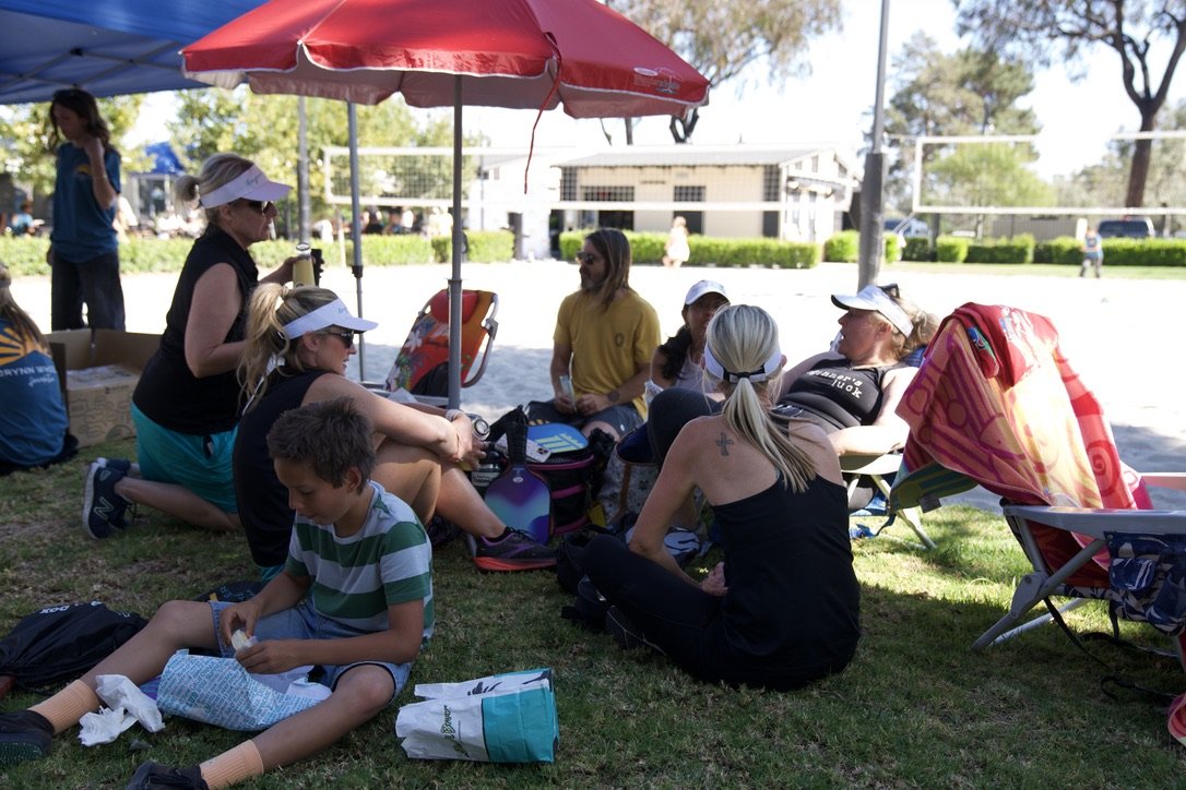 Group of women and children sitting on grass under umbrella at outdoor volleyball court.