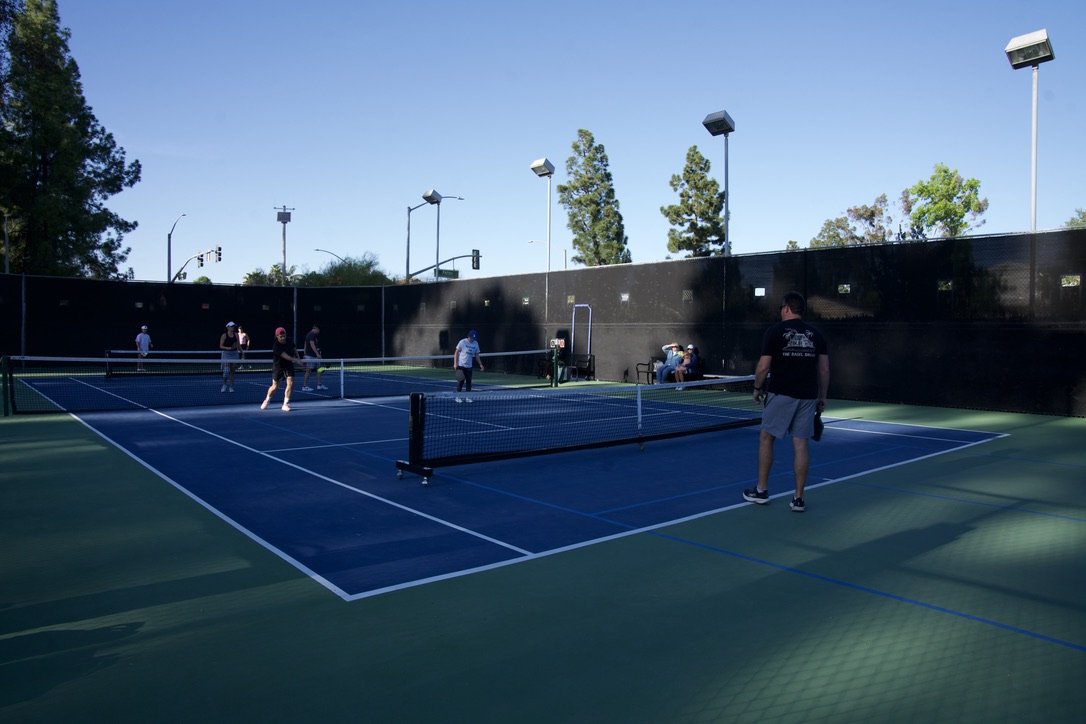 People playing pickleball on an outdoor court with a black windscreen, under a blue sky with trees and tall lights.