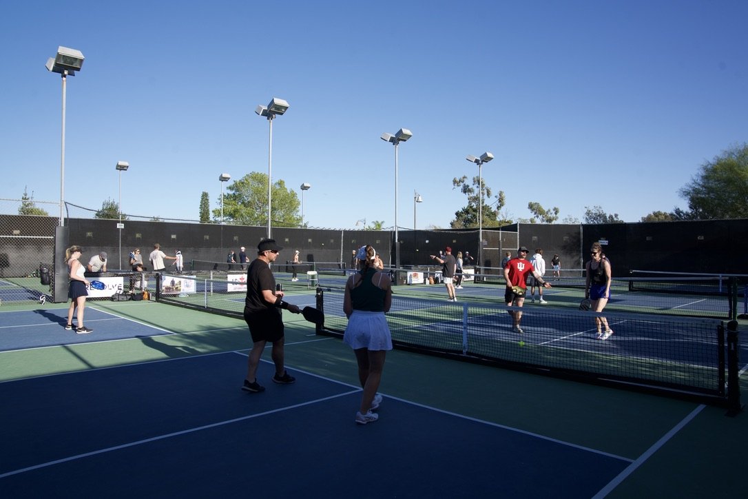 People playing pickleball on multiple outdoor courts during daytime, with some standing near the net and others hitting balls, under a clear blue sky.