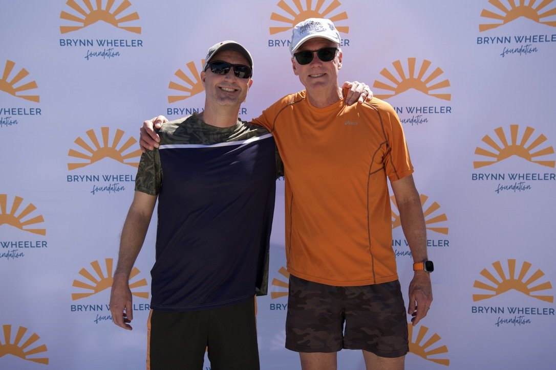 Two men in athletic gear, wearing sunglasses and hats, smiling, with an orange and white backdrop featuring the Brynn Wheeler Foundation logo.