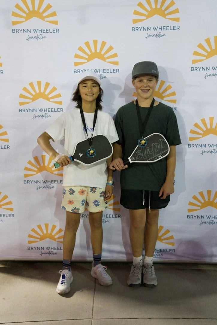 Two kids standing in front of a backdrop with the Brynn Wheeler Foundation logo. The girl on the left is wearing a white T-shirt, colorful shorts, a cap, and holding a paddle with a medal. The boy on the right is wearing a dark T-shirt, shorts, a cap
