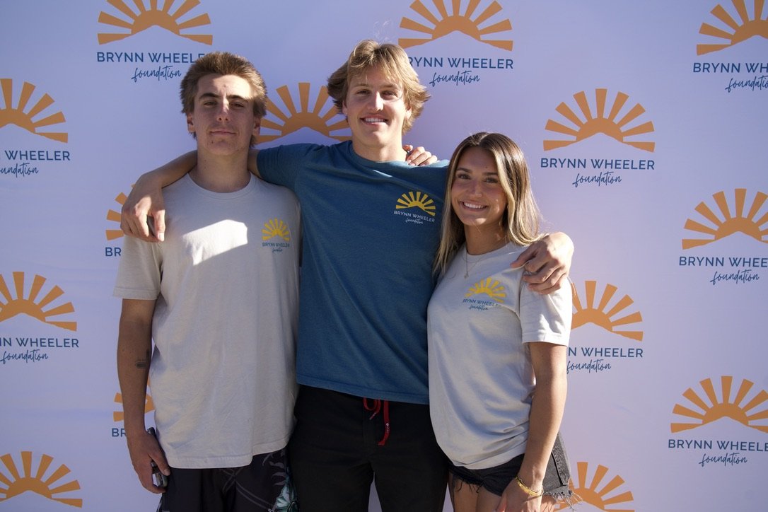 Three young people standing together outdoors, smiling, with a backdrop that has a logo reading 'Brynn Wheeler Foundation' and a yellow sun design. They are wearing t-shirts with the same logo.