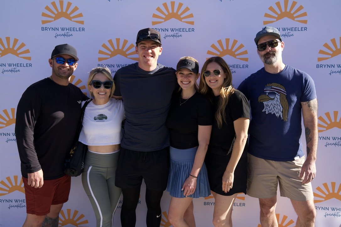 Group of six people smiling and posing in front of a white backdrop with 'Brynn Wheeler Foundation' logo, outdoors on a sunny day.