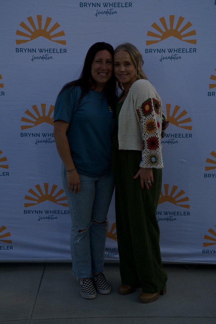Two women standing close together and smiling in front of a backdrop with the Brynn Wheeler Foundation logo, which features a stylized sun with rays.