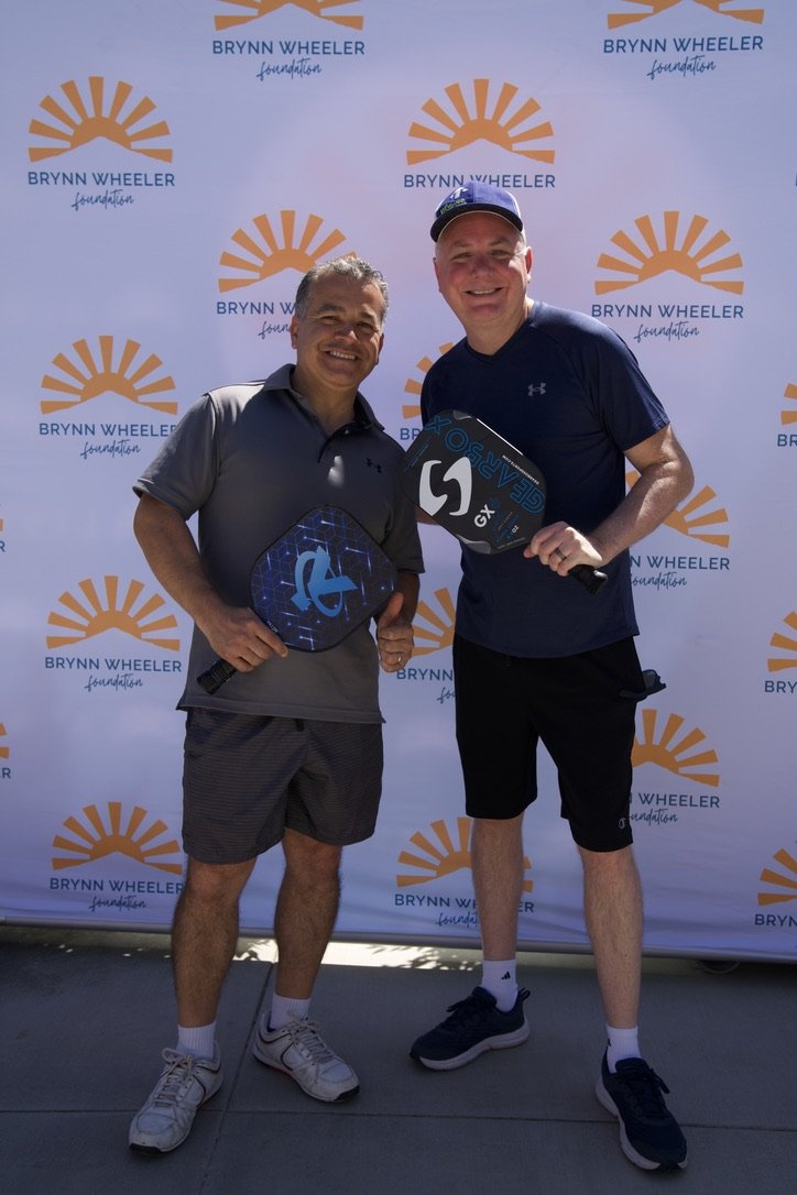 Two men standing in front of a backdrop with the Brynn Wheeler Foundation logo, smiling and holding pickleball paddles.