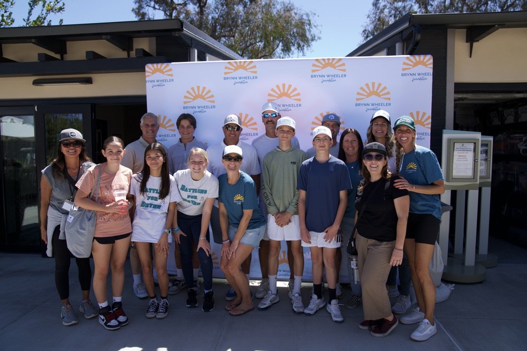 Group of people posing in front of a backdrop with the Brynn Wheeler Foundation logo outside a building on a sunny day.