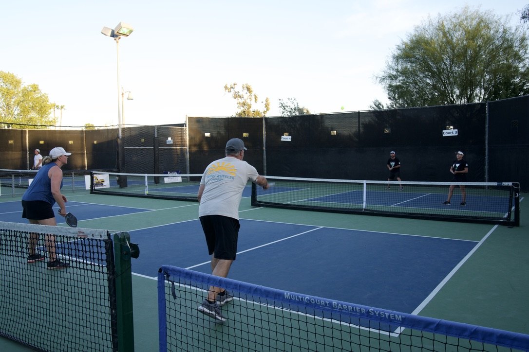 People playing pickleball on an outdoor court during daytime, with black privacy screens and trees in the background.