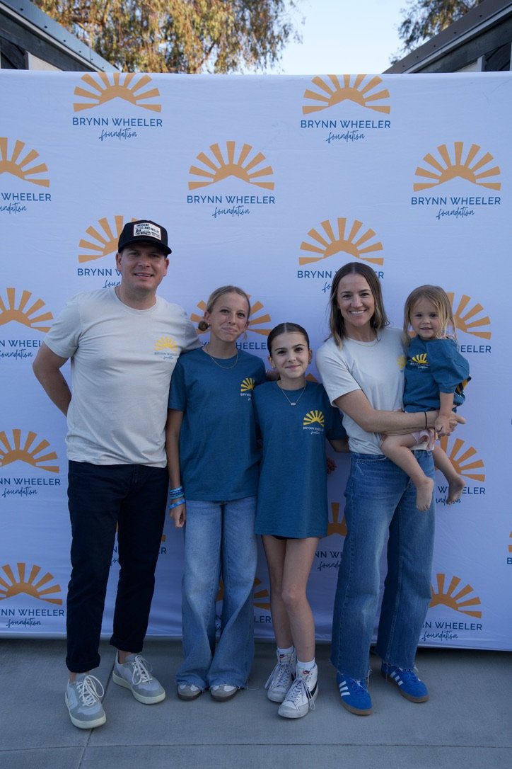 Group of five people, two adults and three children, standing in front of a backdrop with the Brynn Wheeler Foundation logo, outdoors during the daytime.