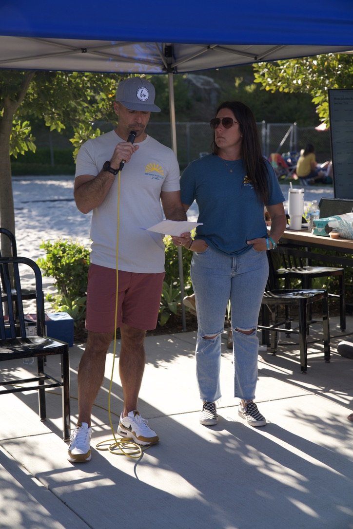 A man with a gray baseball cap, white t-shirt, pink shorts, and sneakers speaking into a microphone while holding a paper, standing next to a woman with long dark hair, sunglasses, jeans, and a blue t-shirt, under a blue canopy outdoors near a lake, 