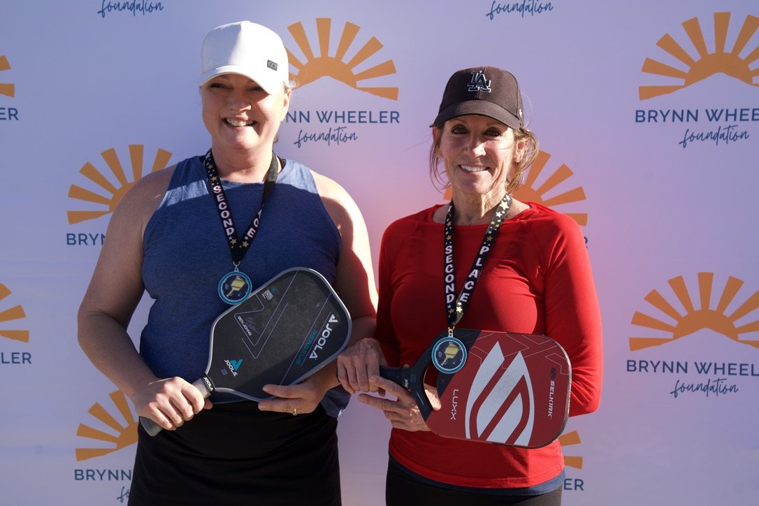 Two women smiling and holding pickleball paddles in front of a Brynn Wheeler Foundation backdrop.