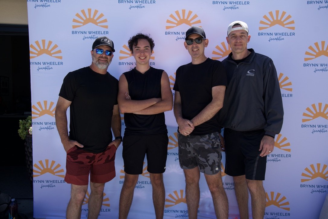 Four people standing in front of a Brynn Wheeler Foundation backdrop. They are dressed casually, likely participating in a sports or outdoor event, and are smiling at the camera.