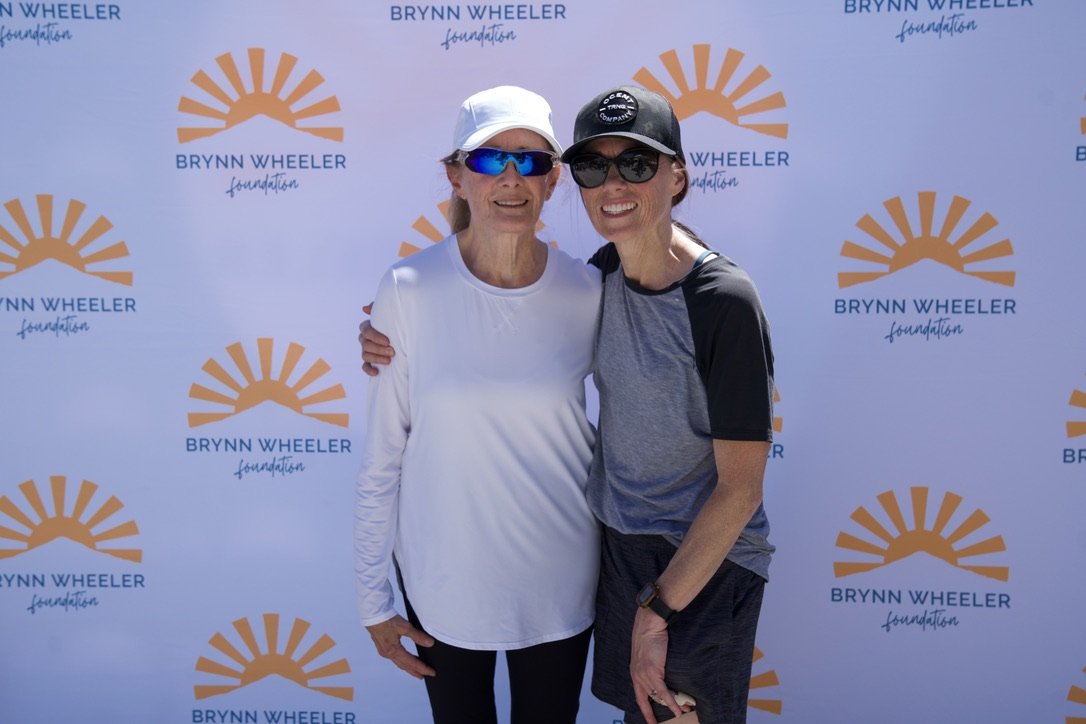 Two women standing together, smiling, at a Brynn Wheeler Foundation event. They are wearing casual athletic clothing and sunglasses, with a backdrop displaying the foundation's logo and name.