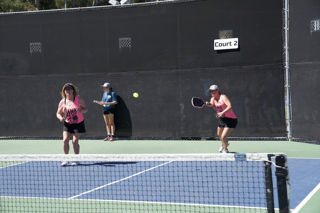 Two women playing pickleball on an outdoor court, with a court sign labeled 'Court 2' in the background. One woman is holding a paddle, and a yellow ball is in mid-air.