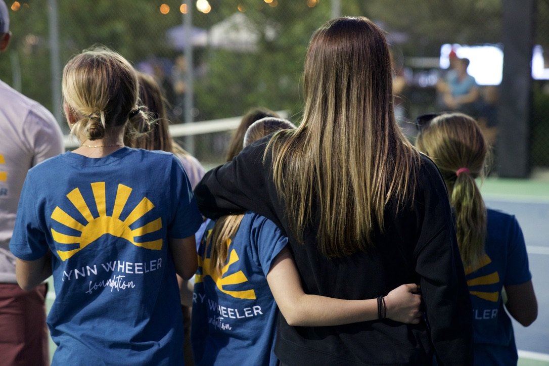 Group of young girls, some wearing blue t-shirts with a sun and mountain logo and the words 'Gwyn Wheeler Foundation,' standing outdoors on a tennis court, with a woman or older girl in black embracing one of the children.
