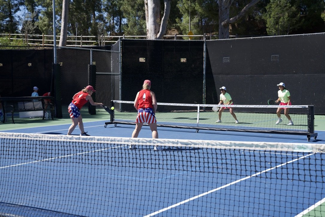 Four women playing pickleball on a blue outdoor court, two on each side of the net, with two behind the net and two in front, surrounded by trees and a black fence.