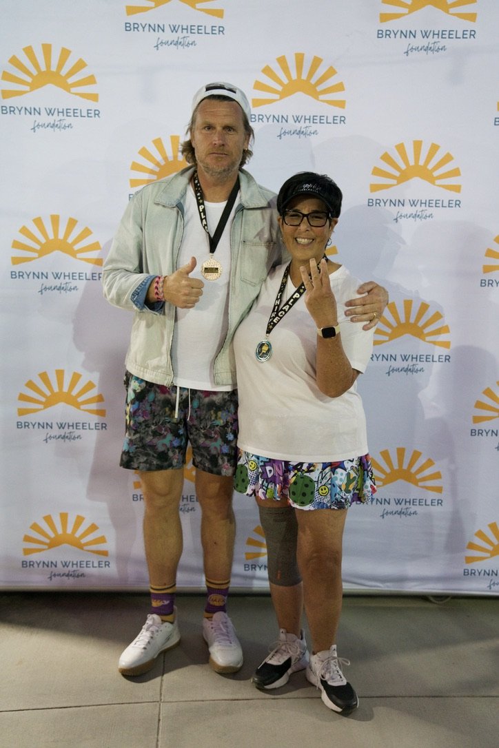 Two people standing together at a Brynn Wheeler Foundation event. The person on the left is a man wearing a white cap backwards, light jacket, white shirt, patterned shorts, and white sneakers, giving a thumbs-up. The person on the right is a woman w