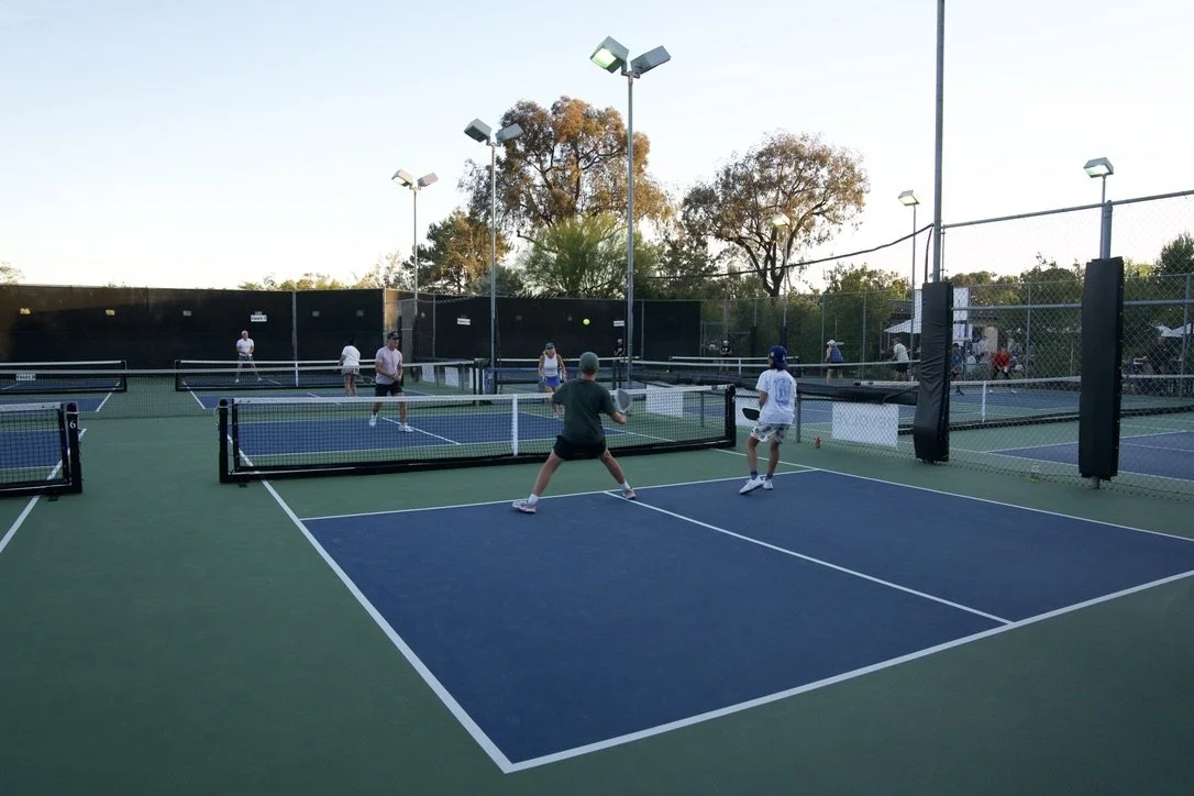 People playing pickleball on outdoor tennis courts with multiple courts, fencing, and trees in the background.