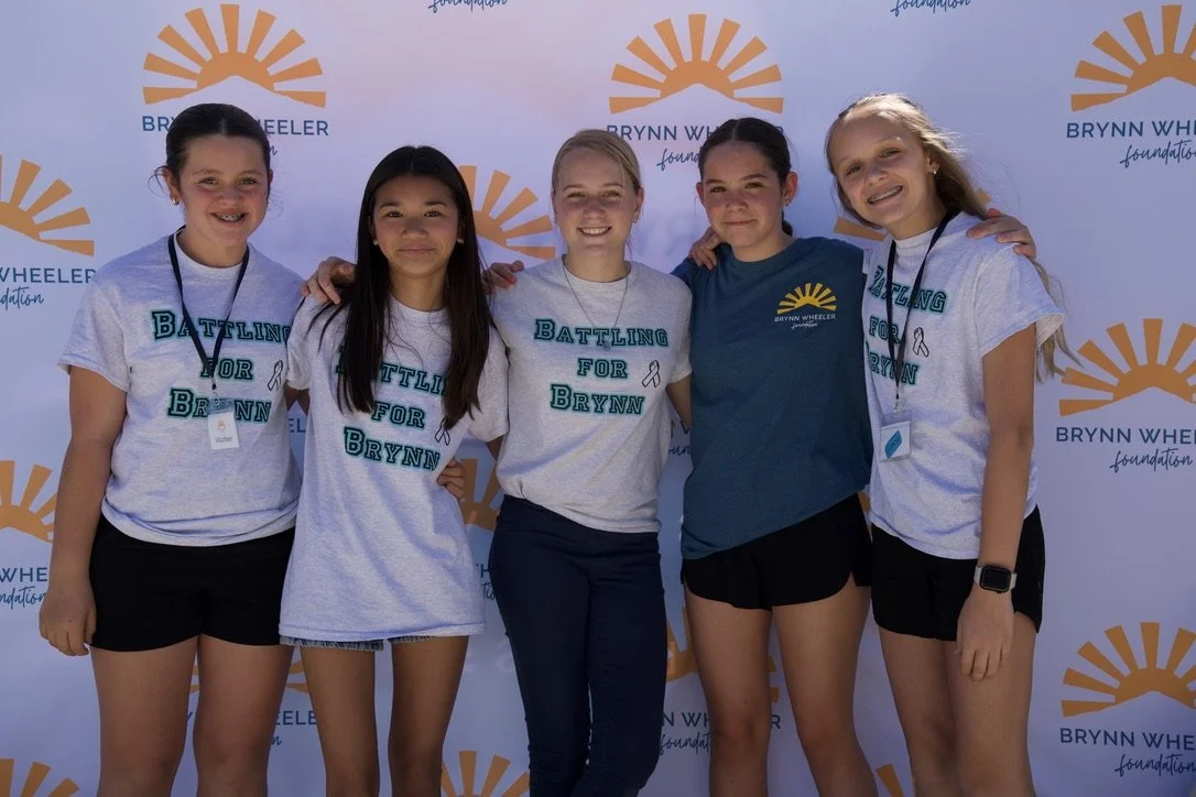 Five young girls standing together in front of a Brynn Wheeler Foundation backdrop, smiling and wearing t-shirts with the slogan 'Battling for Brynn'.