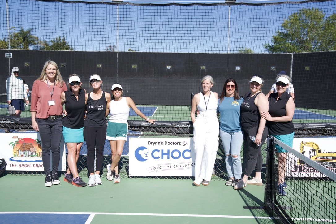 Group of women standing on a tennis court outdoors, smiling for a photo, with tennis nets and trees in the background.