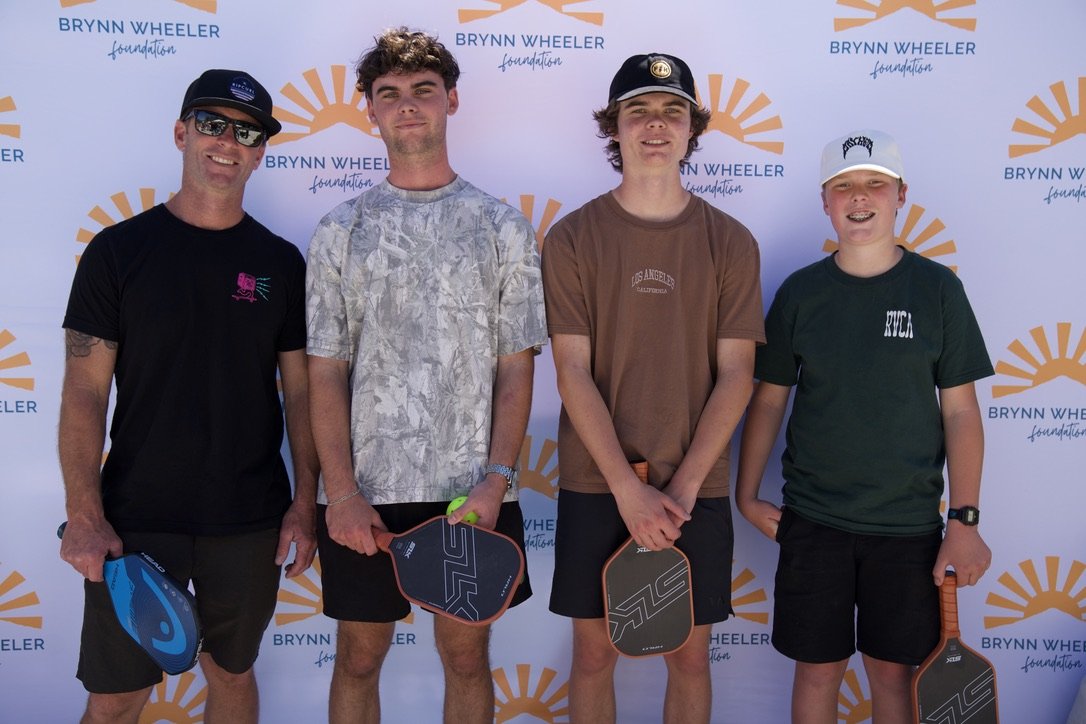 Four young men standing in front of a Brynn Wheeler Foundation backdrop, holding pickleball paddles and smiling at the camera.