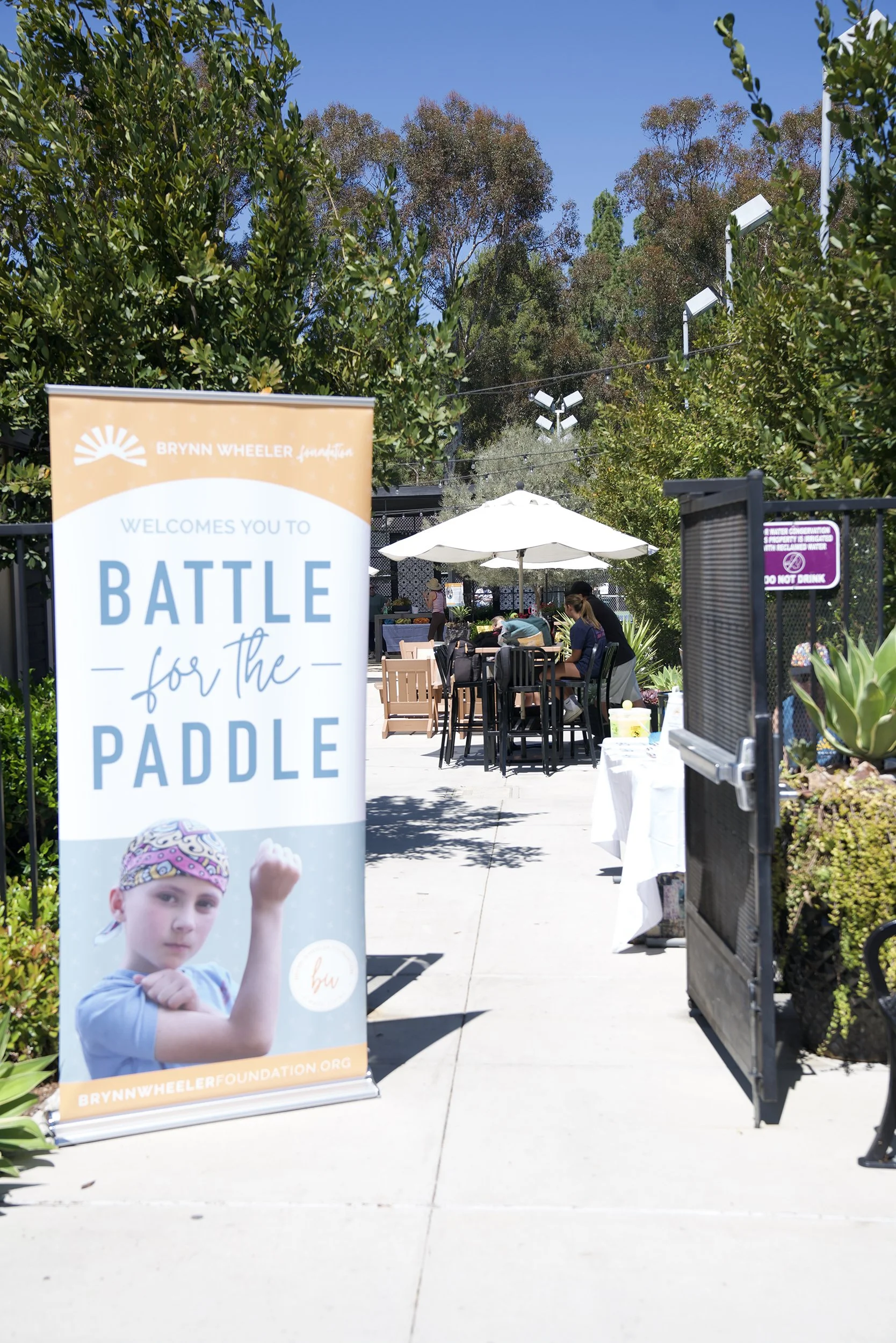 A sign promoting the 'Battle for the Paddle' event by Brynn Wheeler Foundation, with a photo of a young girl in a headband and muscular arm pose, displayed at an outdoor gathering area with people eating under umbrellas and surrounded by trees.