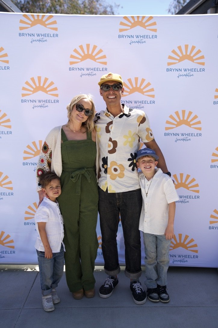 A family of four standing in front of a backdrop with the Brynn Wheeler Foundation logo, which features an orange sunburst and the foundation's name. The family includes a woman with blonde hair wearing sunglasses and a cardigan, a man with dark sung