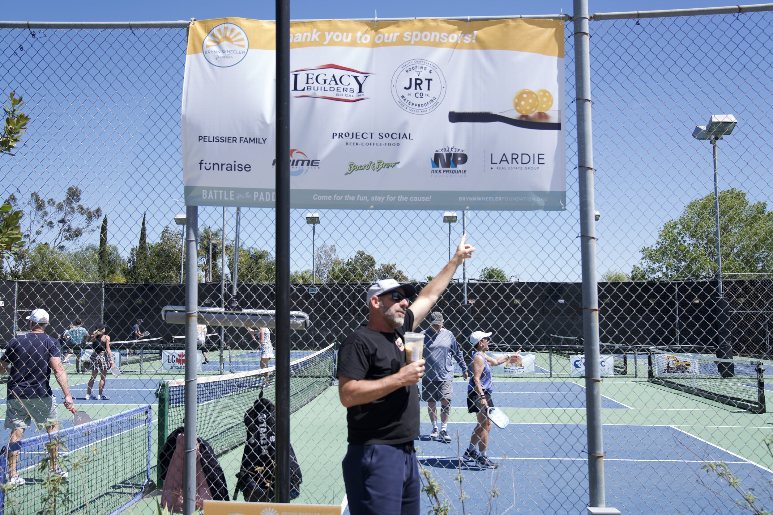 People playing pickleball on courts with a man in black shirt and sunglasses pointing at a large sponsor banner.