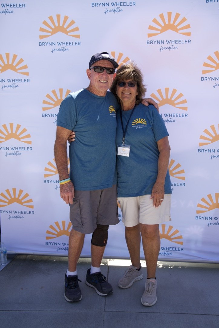 Two people standing together in front of a backdrop with the Brynn Wheeler Foundation logo, both wearing matching blue shirts. They are smiling, with one person's arm around the other's shoulder.