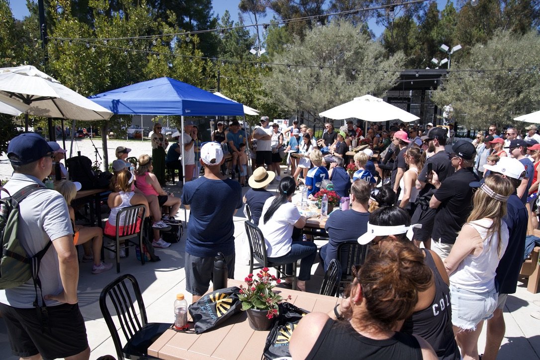 Crowd gathered outdoors for an event on a sunny day, with umbrellas and tents providing shade, surrounded by trees.