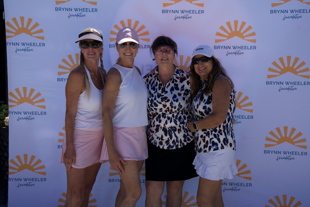 Four women standing in front of a Brynn Wheeler Foundation backdrop, dressed in sporty outfits, wearing visors and sunglasses, smiling at the camera.