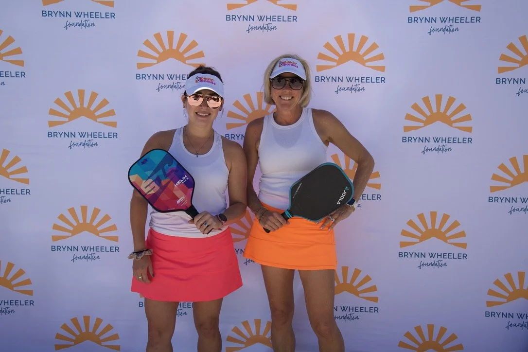 Two women in athletic outfits holding paddles, smiling, standing in front of a Brenda Wheeler Foundation backdrop.