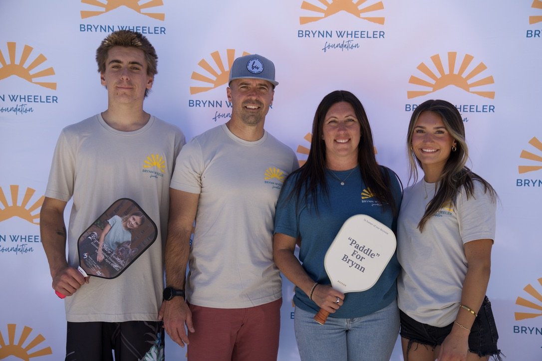 Four people standing in front of a backdrop with the Brynn Wheeler Foundation logo. The group includes two men and two women, all smiling, with two holding fans that say "Paddle For Brynn."