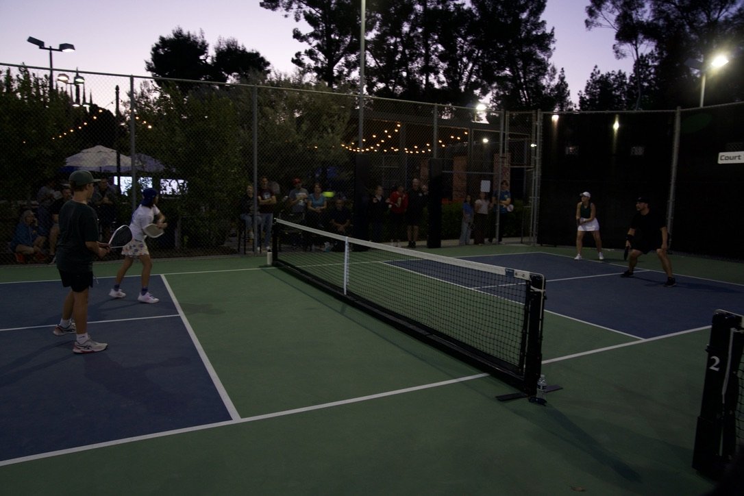 People playing doubles tennis on an outdoor court at dusk, with a group of spectators watching behind a fence.