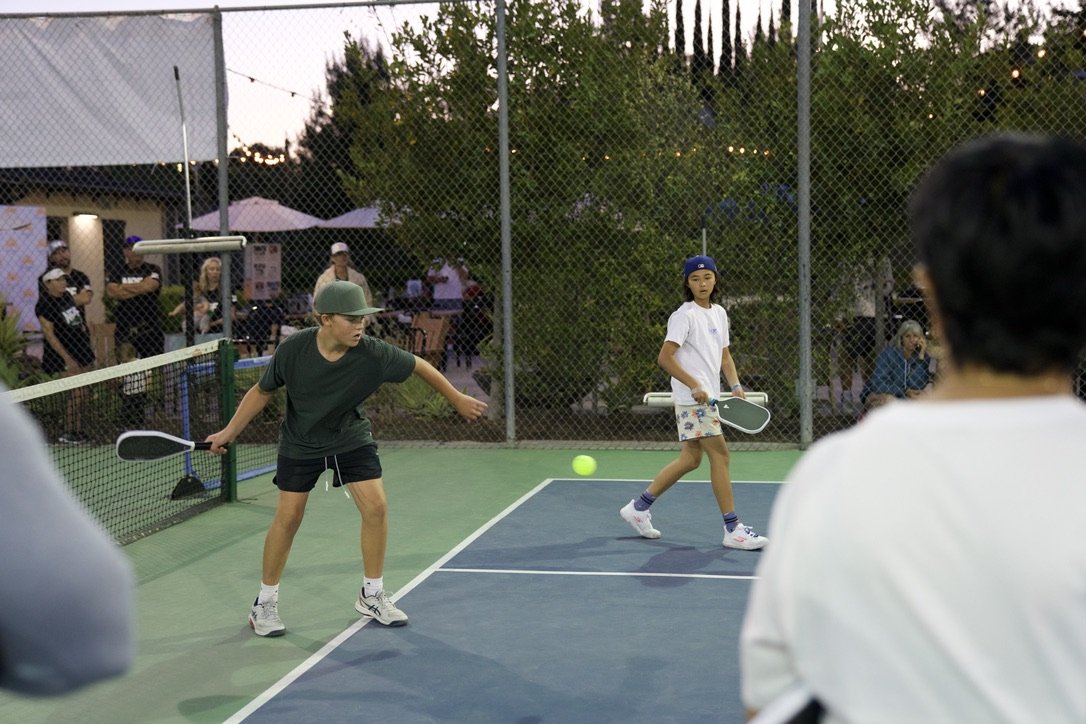 Two children playing pickleball on an outdoor court during the evening. One boy is wearing a green cap, a dark t-shirt, shorts, and sneakers, and is hitting a yellow ball with a paddle. A girl in a blue cap, white t-shirt, colorful shorts, and sneake