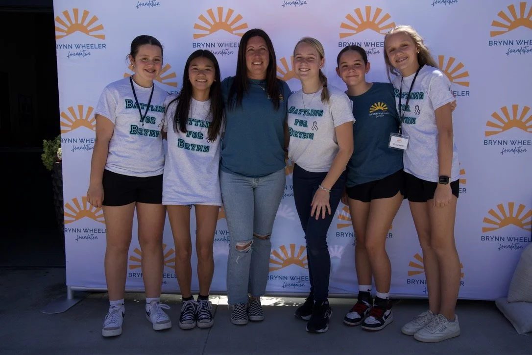 Six girls and one woman standing in front of a Brynn Wheeler Foundation backdrop, smiling for a group photo at an outdoor event.