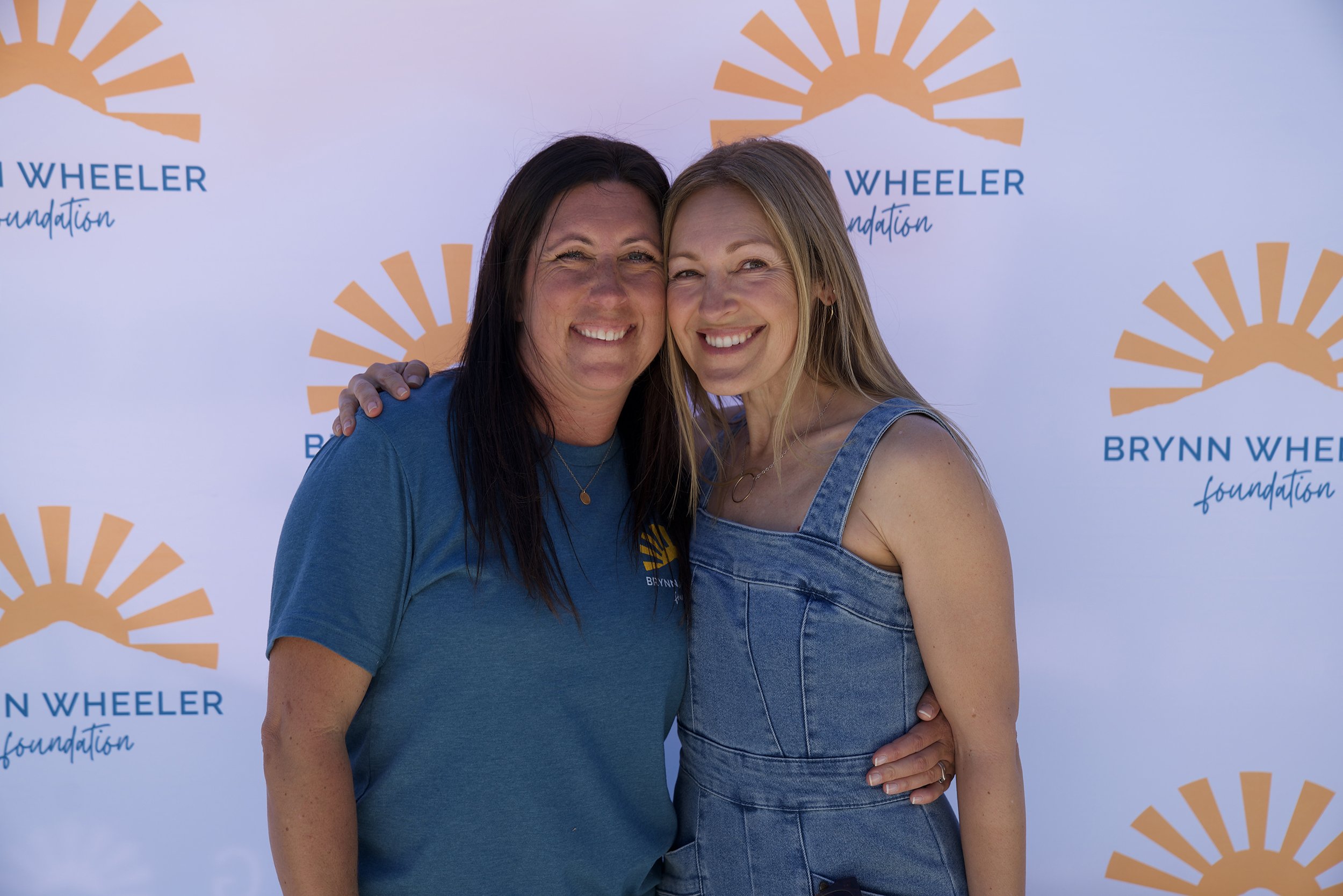 Two women standing close together, smiling, in front of a backdrop with the Brynn Wheeler Foundation logo. The woman on the left has dark hair, and the woman on the right has blonde hair, and is wearing a denim sleeveless top.