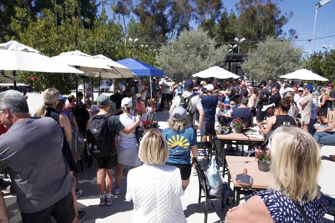 Crowd gathering outdoors at a shaded event with tables, chairs, umbrellas, and string lights.