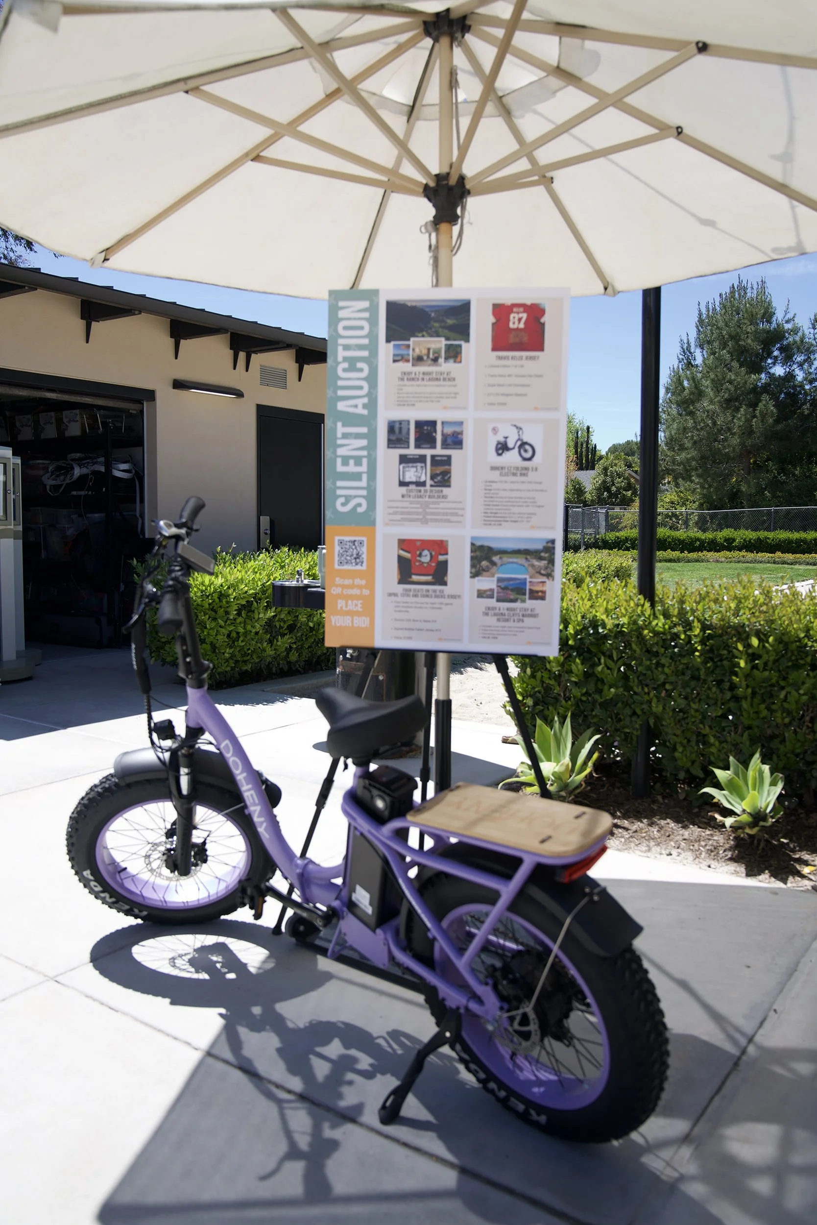 Purple electric bicycle parked outside under a large cream-colored patio umbrella near a silent auction sign with information, images, and a QR code. Formation of trees and a building are visible in the background.