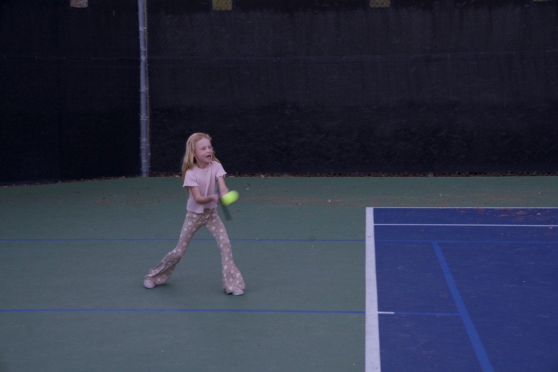Young girl playing tennis on an outdoor court, holding a tennis racket with a tennis ball in her hand.