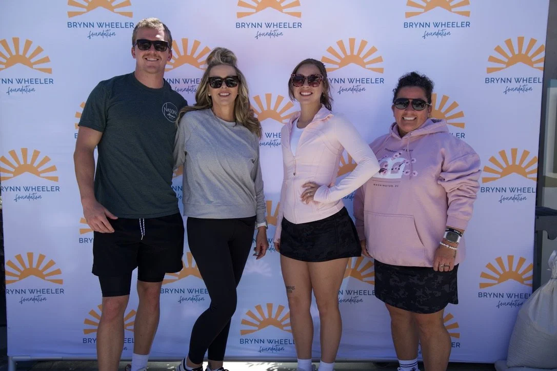 Four people smiling and posing in front of a Brynn Wheeler Foundation backdrop. They are wearing sunglasses and casual clothing.