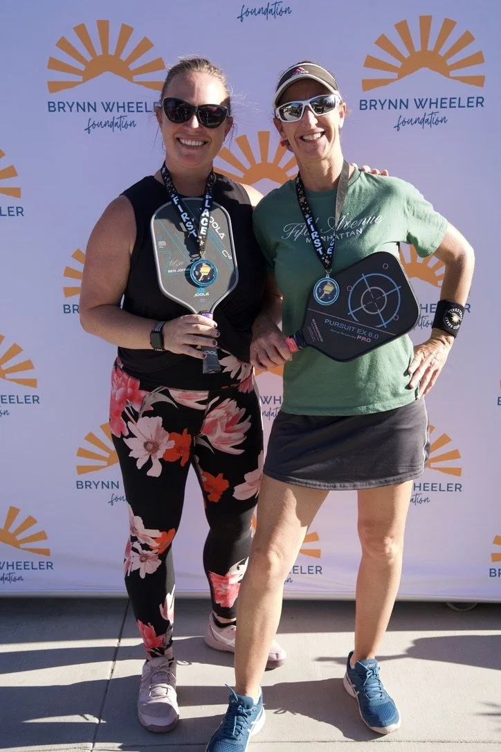 Two women standing side by side, smiling and holding medals and paddleboards, at a Brynn Wheeler Foundation event. One woman is wearing a black tank top and floral leggings, while the other is in a green T-shirt and black skirt. They are outdoors, wi