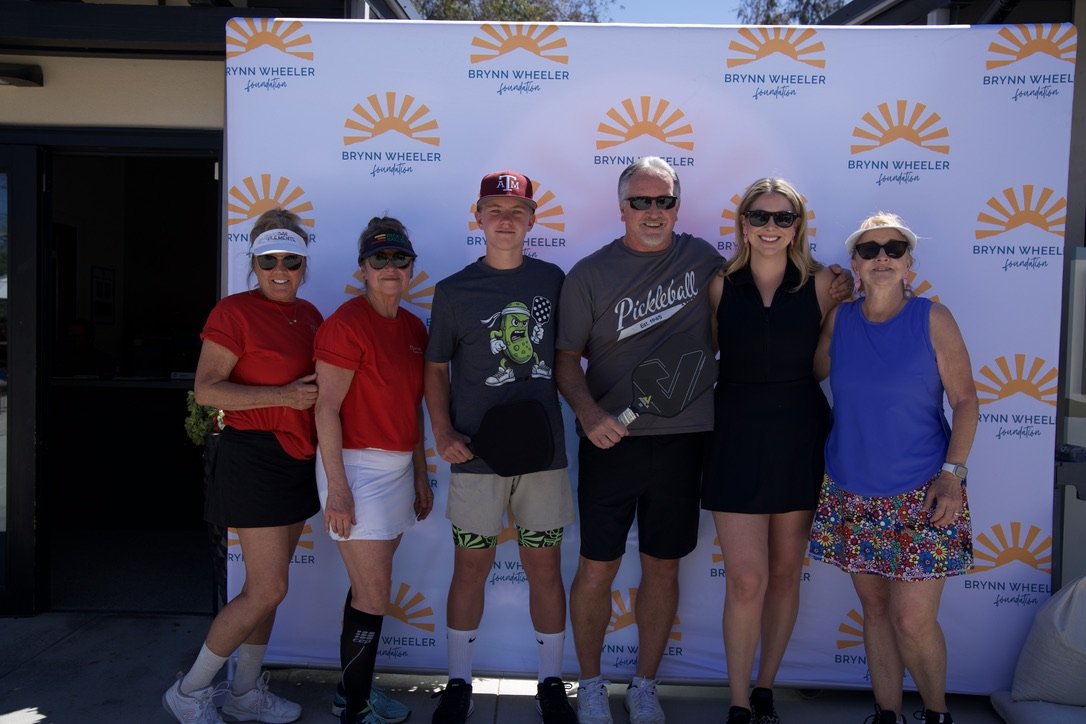 Group of six people standing outdoors in front of a Brynn Wheeler Foundation backdrop, smiling for a photo. The group includes women and men wearing casual attire, some with sunglasses and hats.