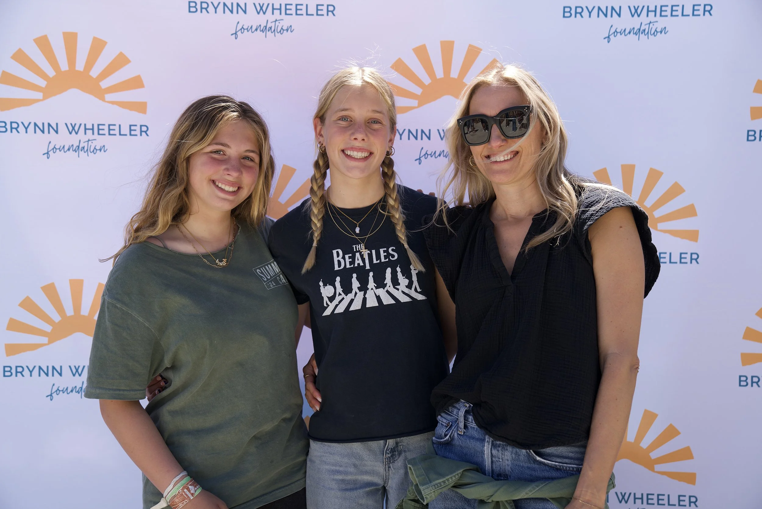 Three women standing together outdoors in front of a backdrop with the Brynn Wheeler Foundation logo, smiling at the camera.