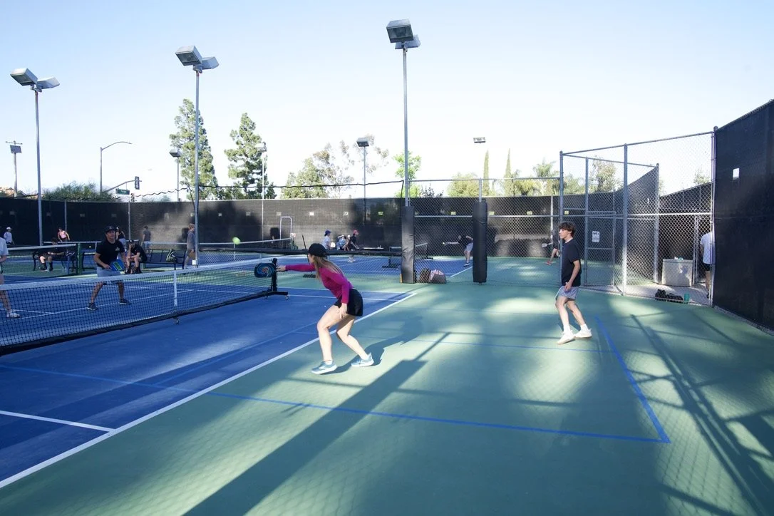 People playing pickleball on outdoor courts with chain-link fencing in a sunny setting.