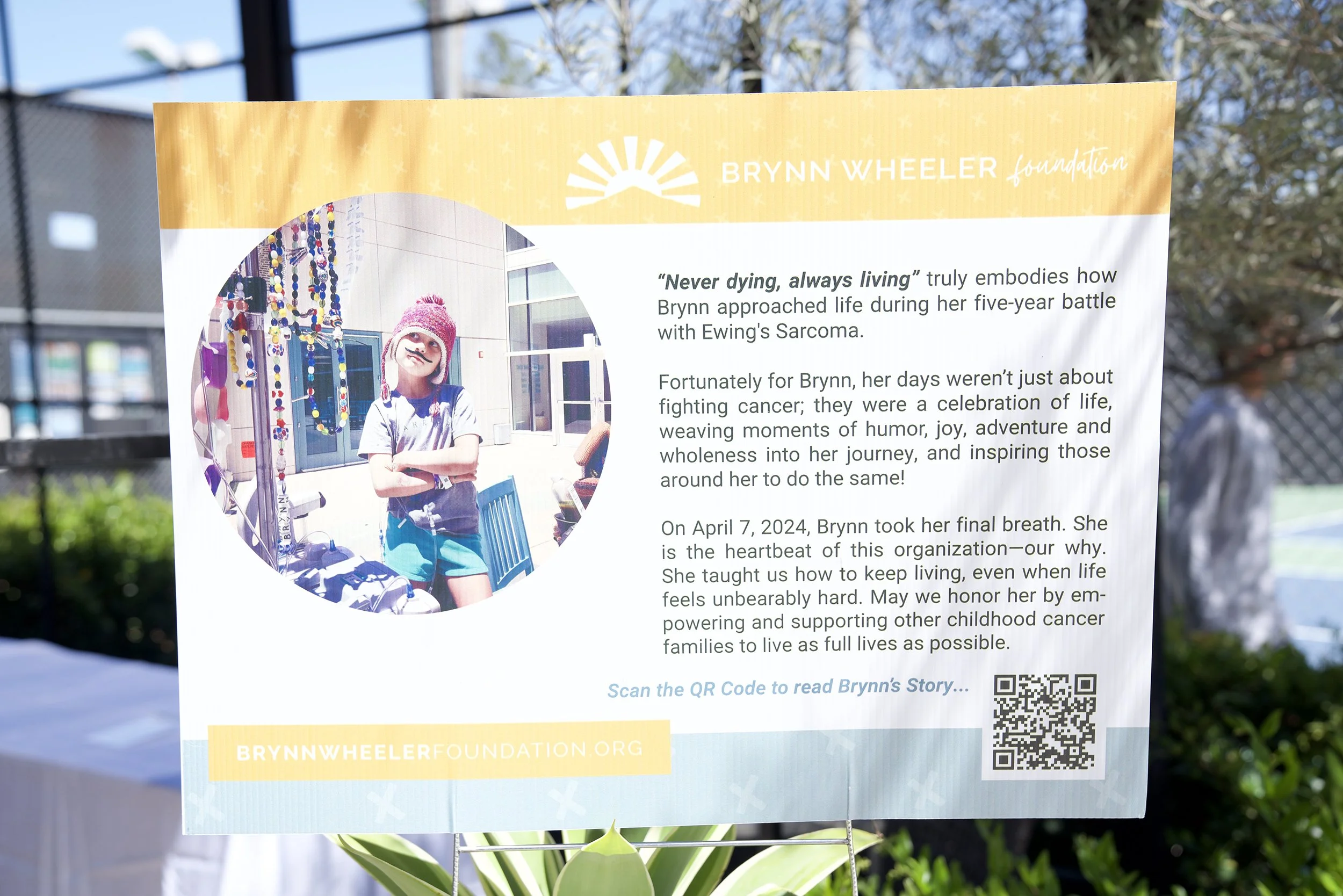 A sign with a photo of Brynn in a pink beanie and grey t-shirt, standing outdoors with arms crossed, featuring yellow and white text about her battle with Ewing's Sarcoma and her legacy, including a QR code and website link.