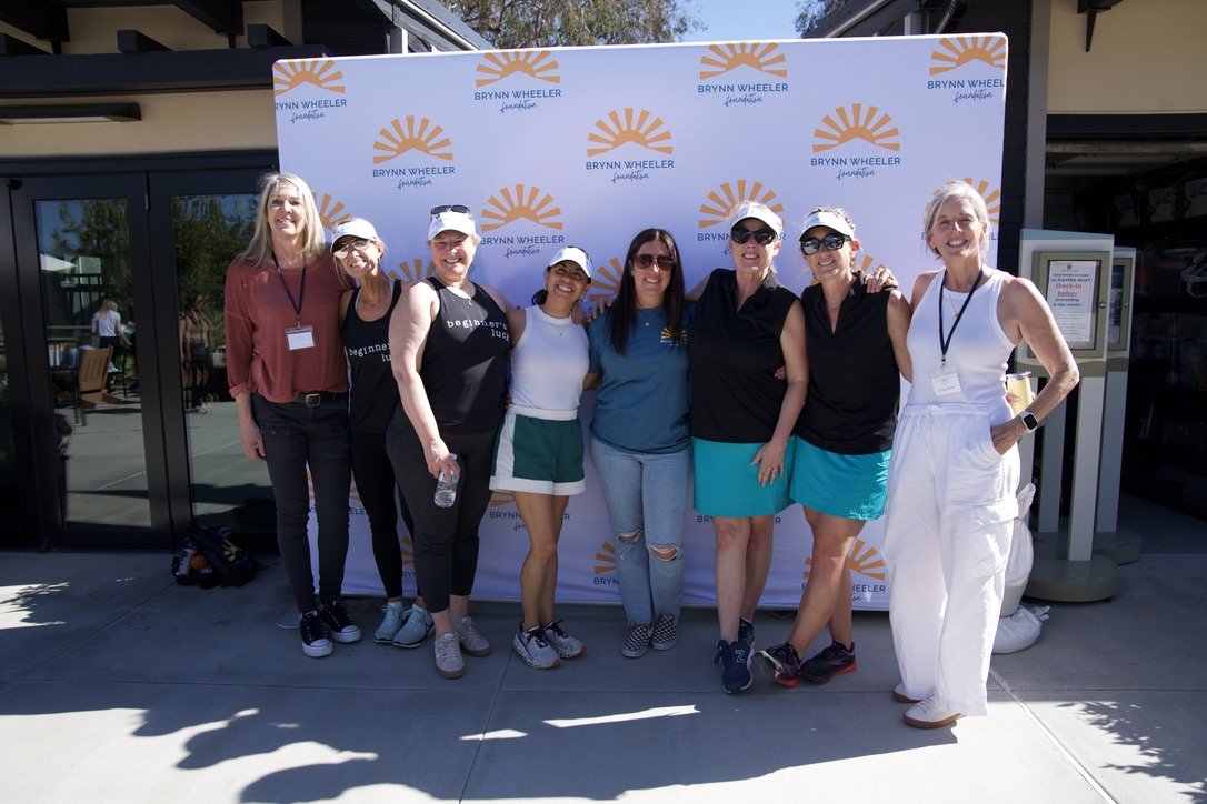 Group of eight women standing in front of a Brynn Wheeler Foundation backdrop, smiling outdoors.