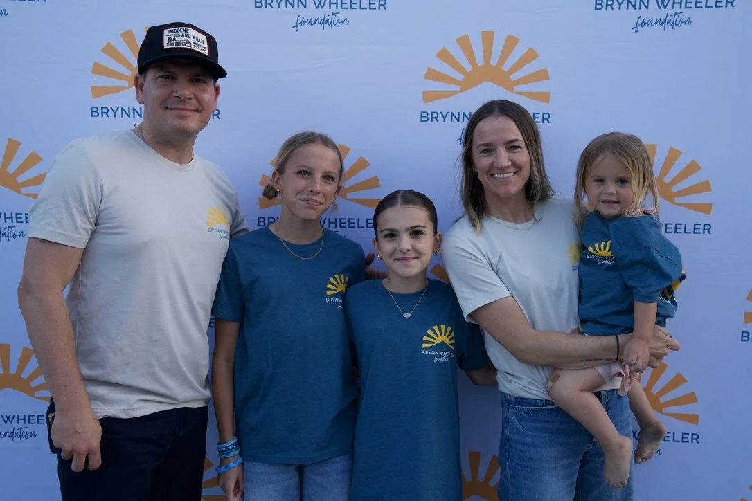 A family standing in front of a Brynn Wheeler Foundation backdrop, wearing matching T-shirts with a sun logo, smiling at the camera.
