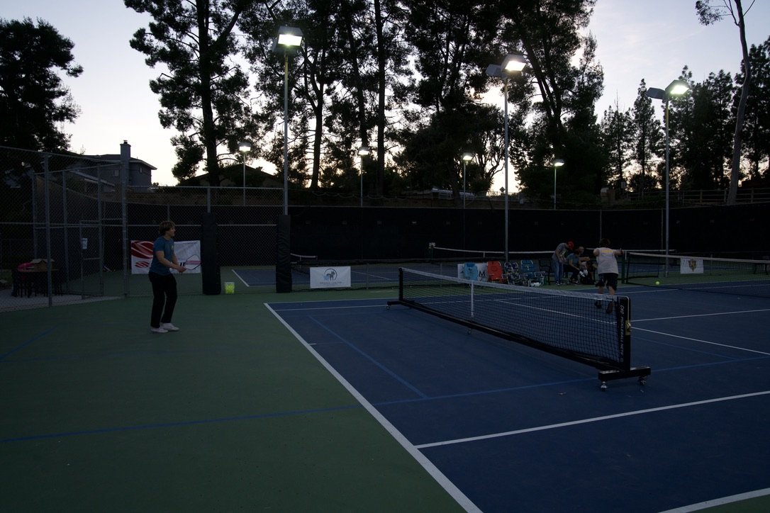 People playing tennis at an outdoor tennis court during dusk, with tall trees and floodlights illuminating the court.