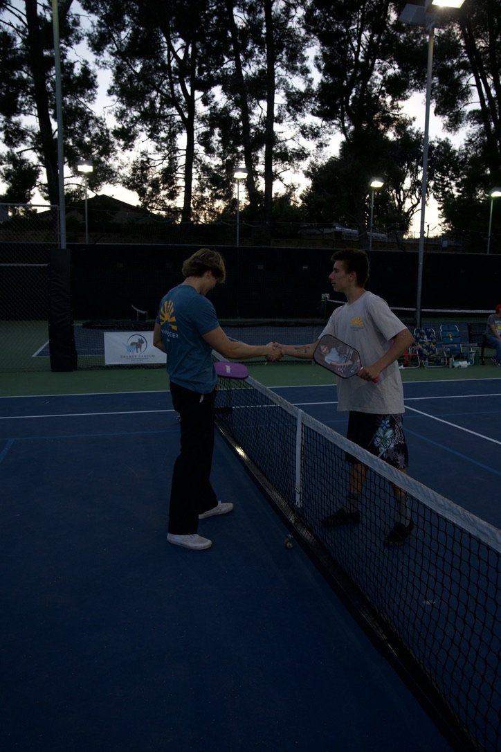 Two young men shake hands after a tennis match on an outdoor court during dusk, with trees and lights in the background.