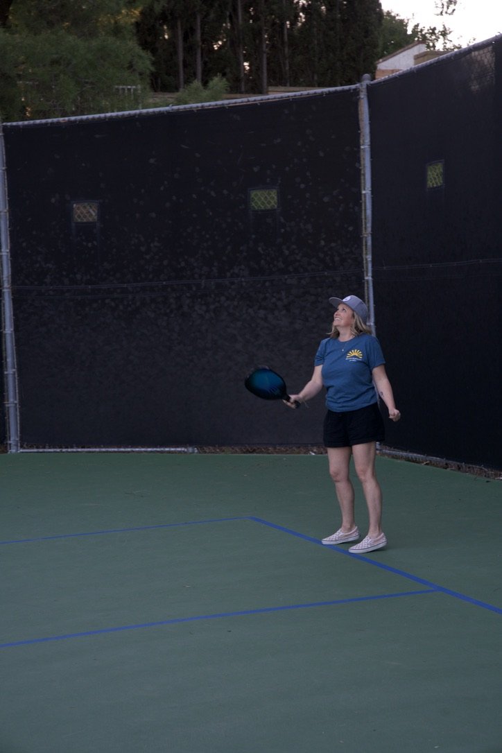 A woman playing pickleball on an outdoor court at dusk, holding a paddle and smiling.